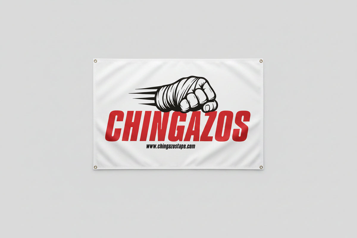 A white rectangular banner hanging on a concrete wall, printed with large red vertical text reading 'CHINGAZOS,' a black-and-white illustrated fist with claws, and small black website text. The banner has metal grommets at the corners.